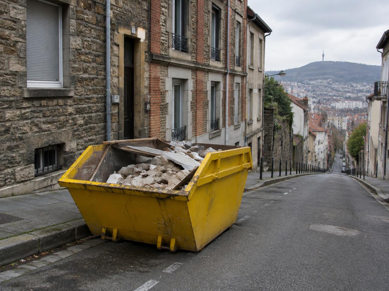 Benne jaune chargée de gravats dans une rue en pente de Saint-Étienne